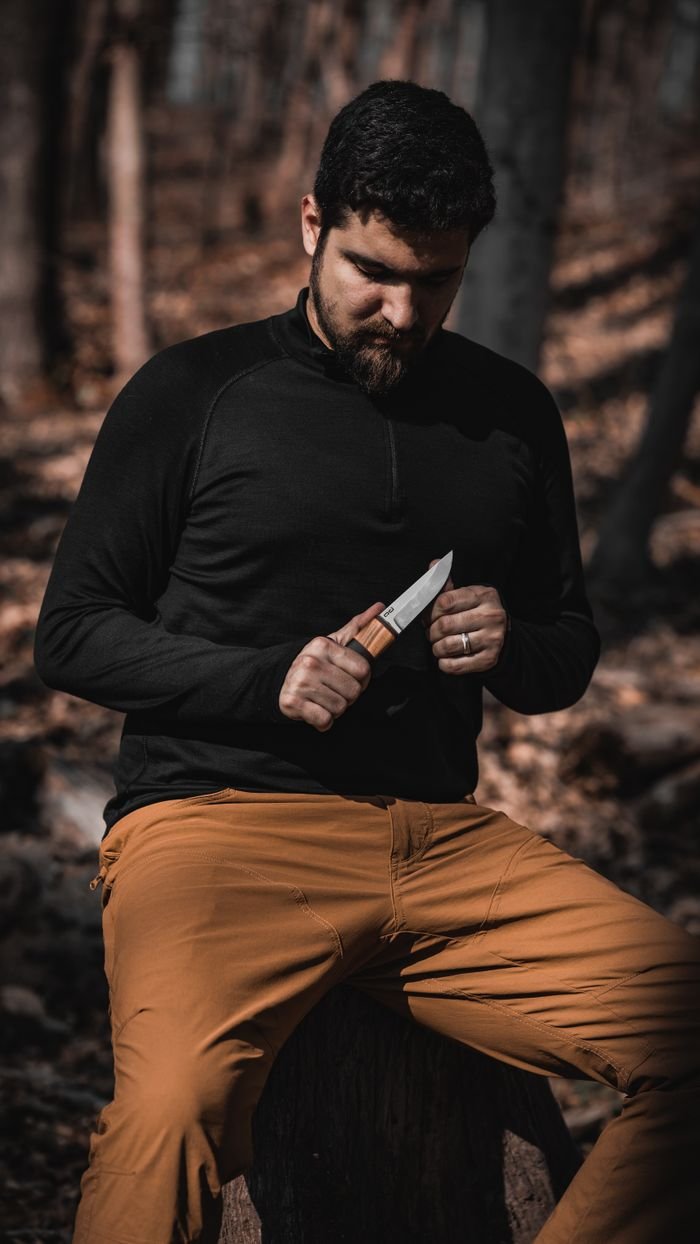 About A man in a forest sitting on a tree stump, holding a knife during a daytime hike.